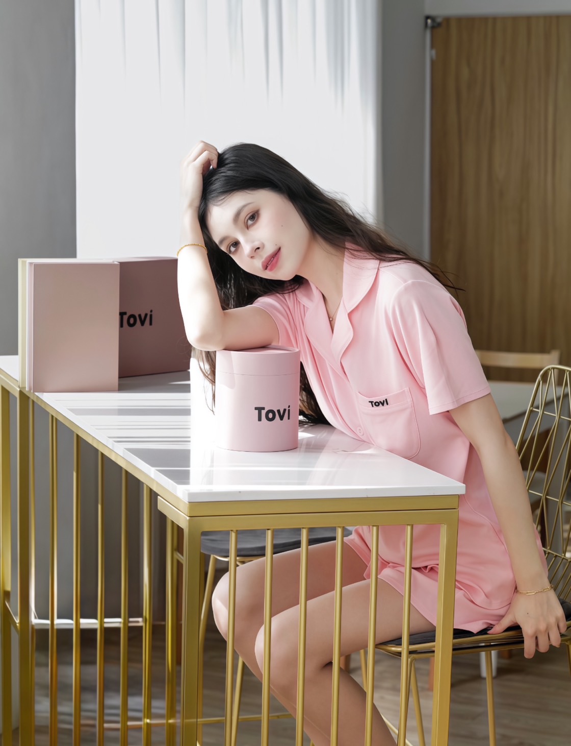 Model in pink Tovi sleepwear resting at a sunlit table with Tovi gift boxes